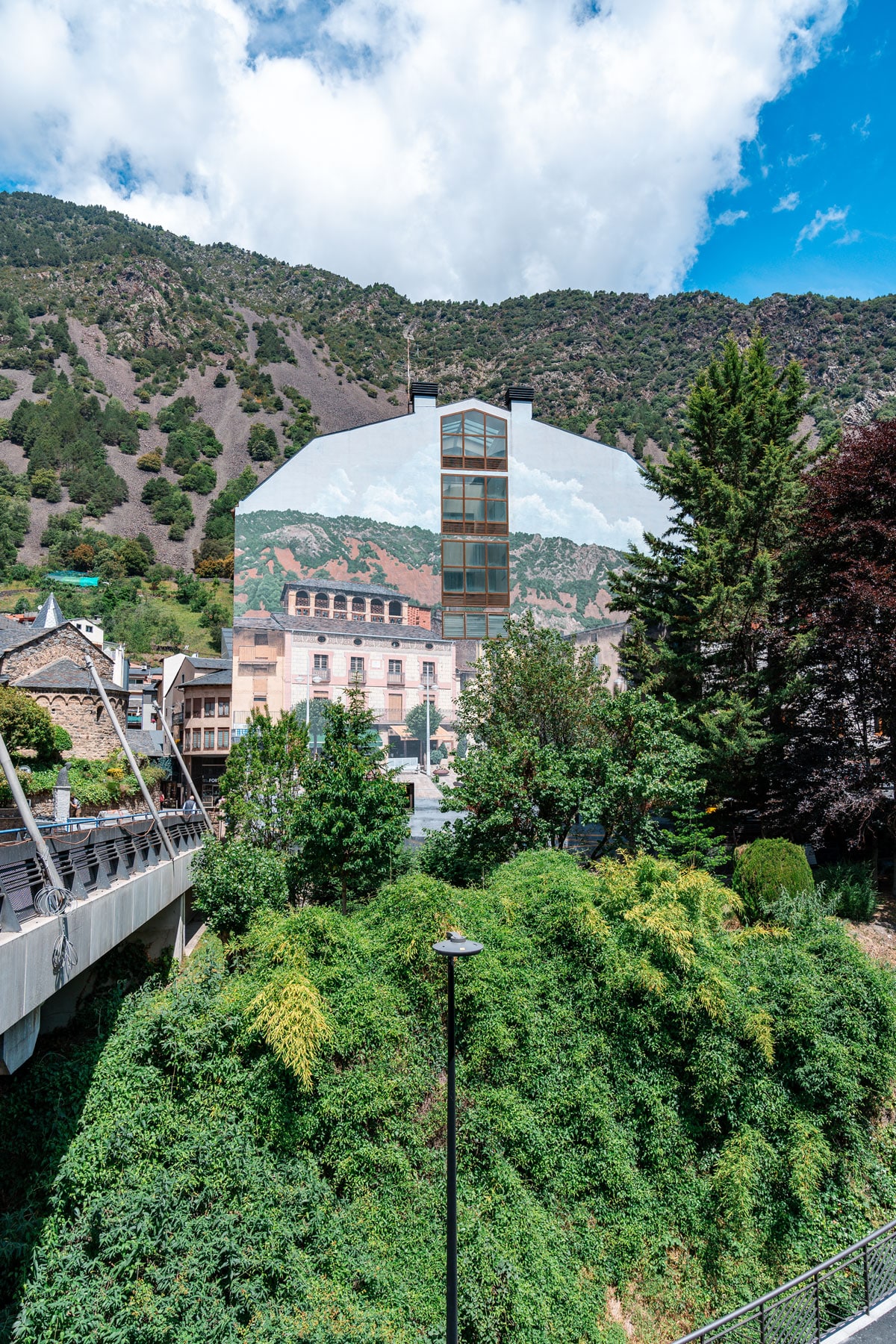 Riverside building covered with a huge mountain mural, backed by real hills in Andorra la Vella.