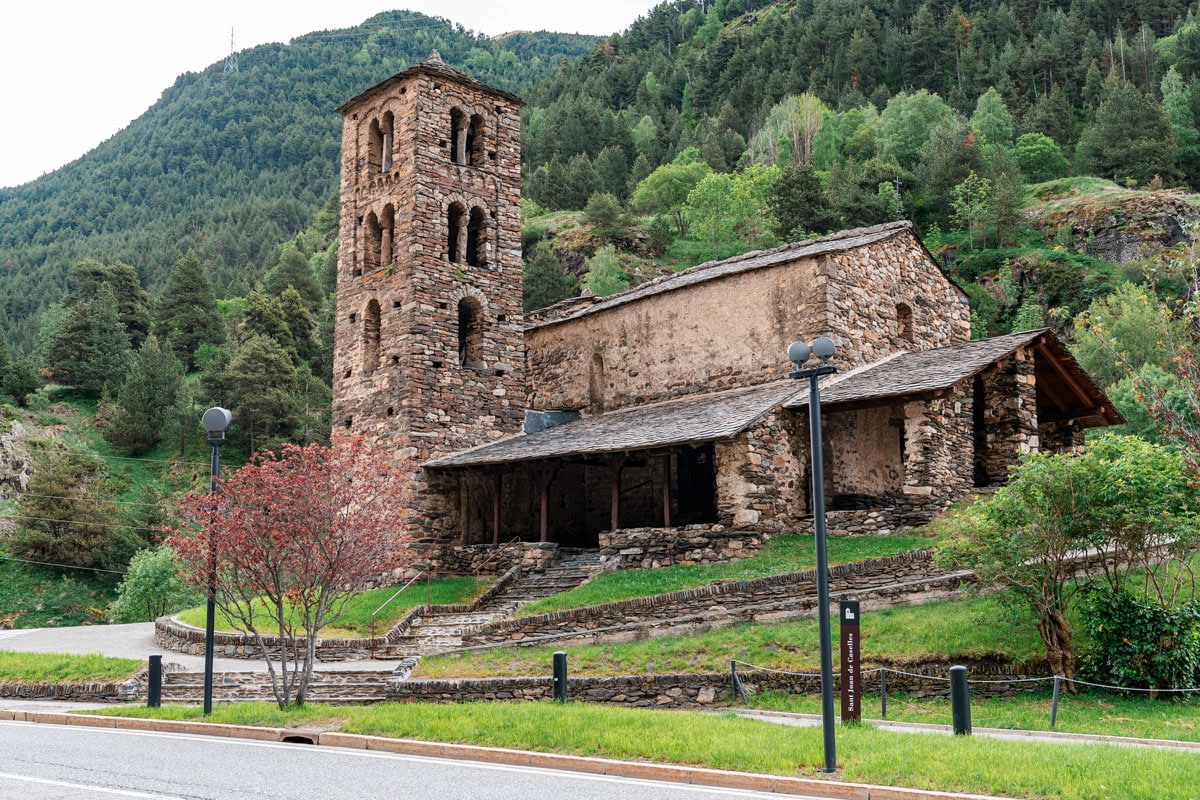Stone Sant Joan de Caselles church and tower beside the road, with forested mountains behind.