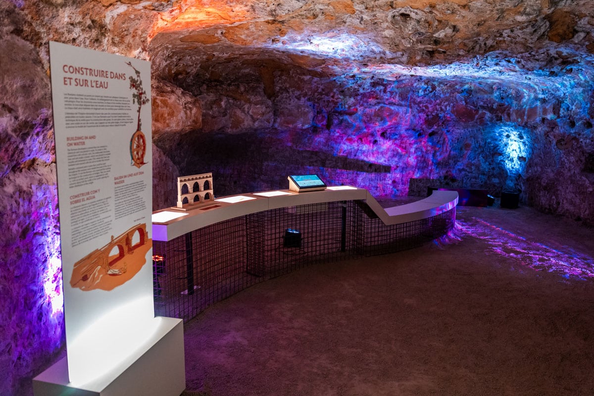 A cave gallery beneath the theatre lit in purple, with a curved walkway and an information panel.