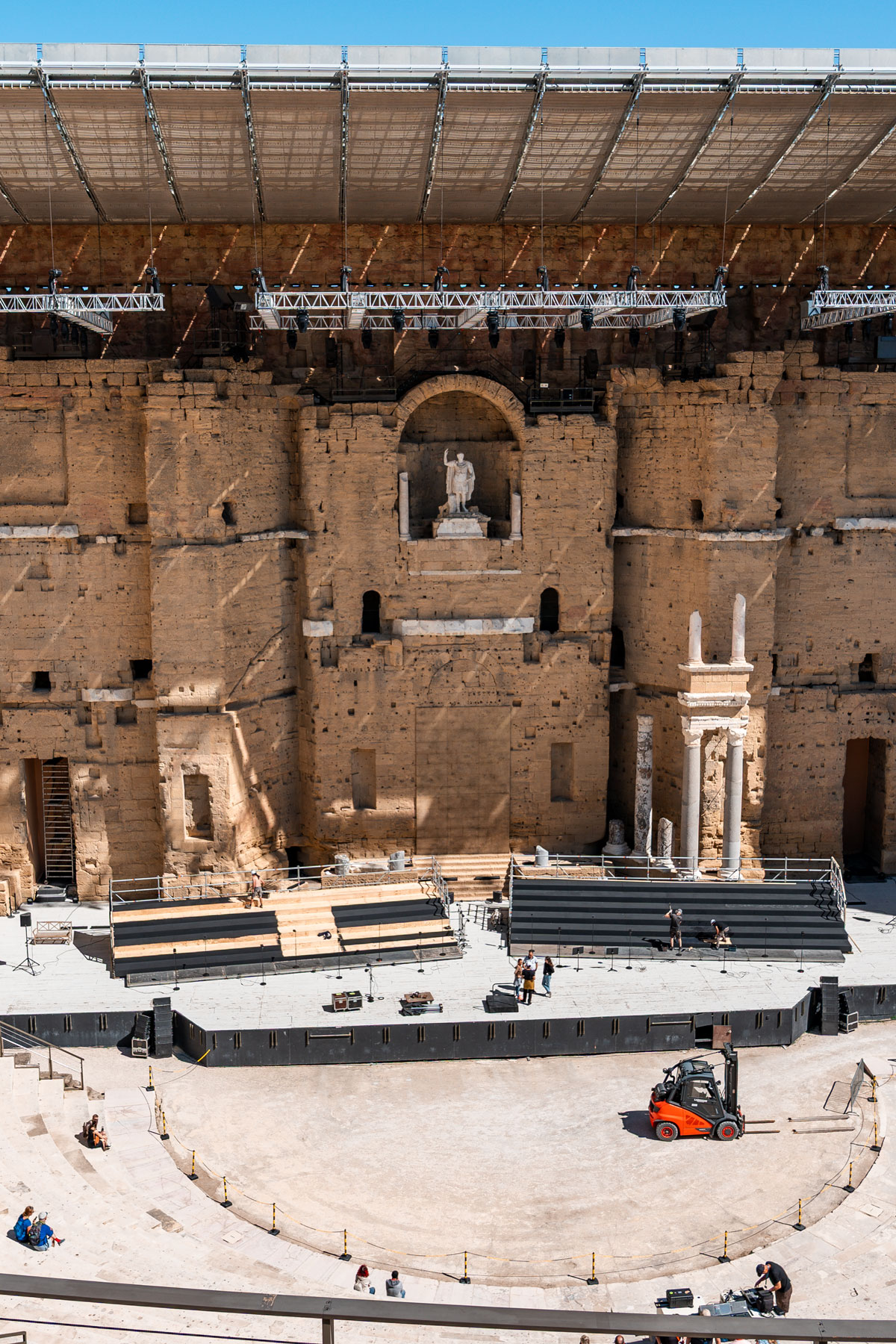 The Roman Theatre of Orange seen from the upper seating, with the tall stone stage wall and curved rows of stone seats under a blue sky.