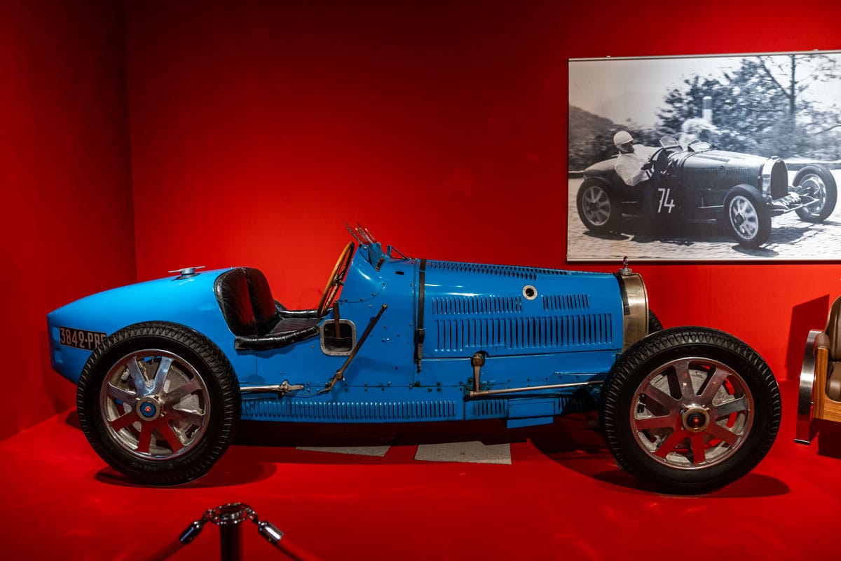 Blue Bugatti racing car in a red-walled room beside a black-and-white racing photograph.