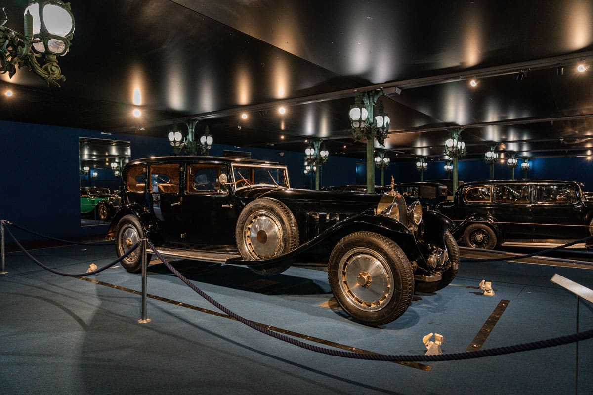 Black Bugatti Royale behind a rope barrier in a dark blue gallery lined with mirrors.