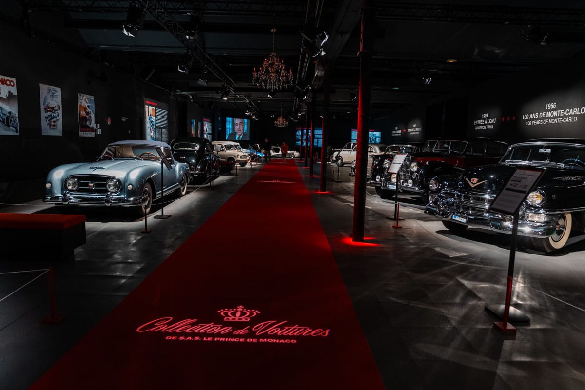 Cars from the Prince of Monaco’s collection lined along a red carpet in a darkened gallery.