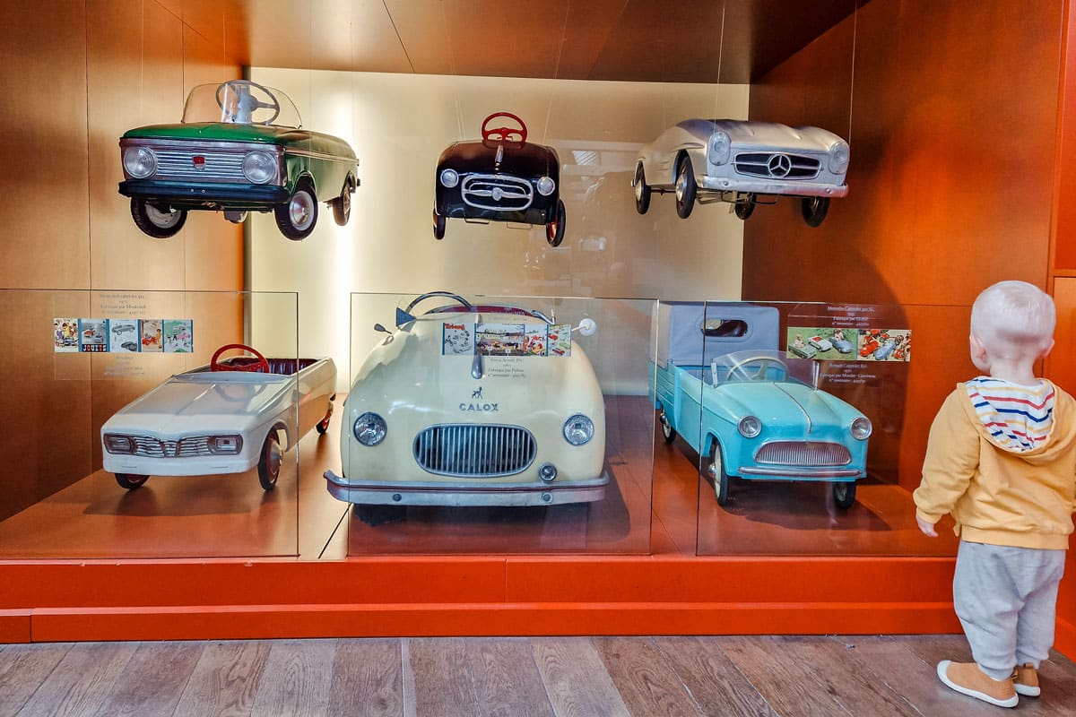 Our son in a yellow hoodie looking at vintage pedal cars behind glass.